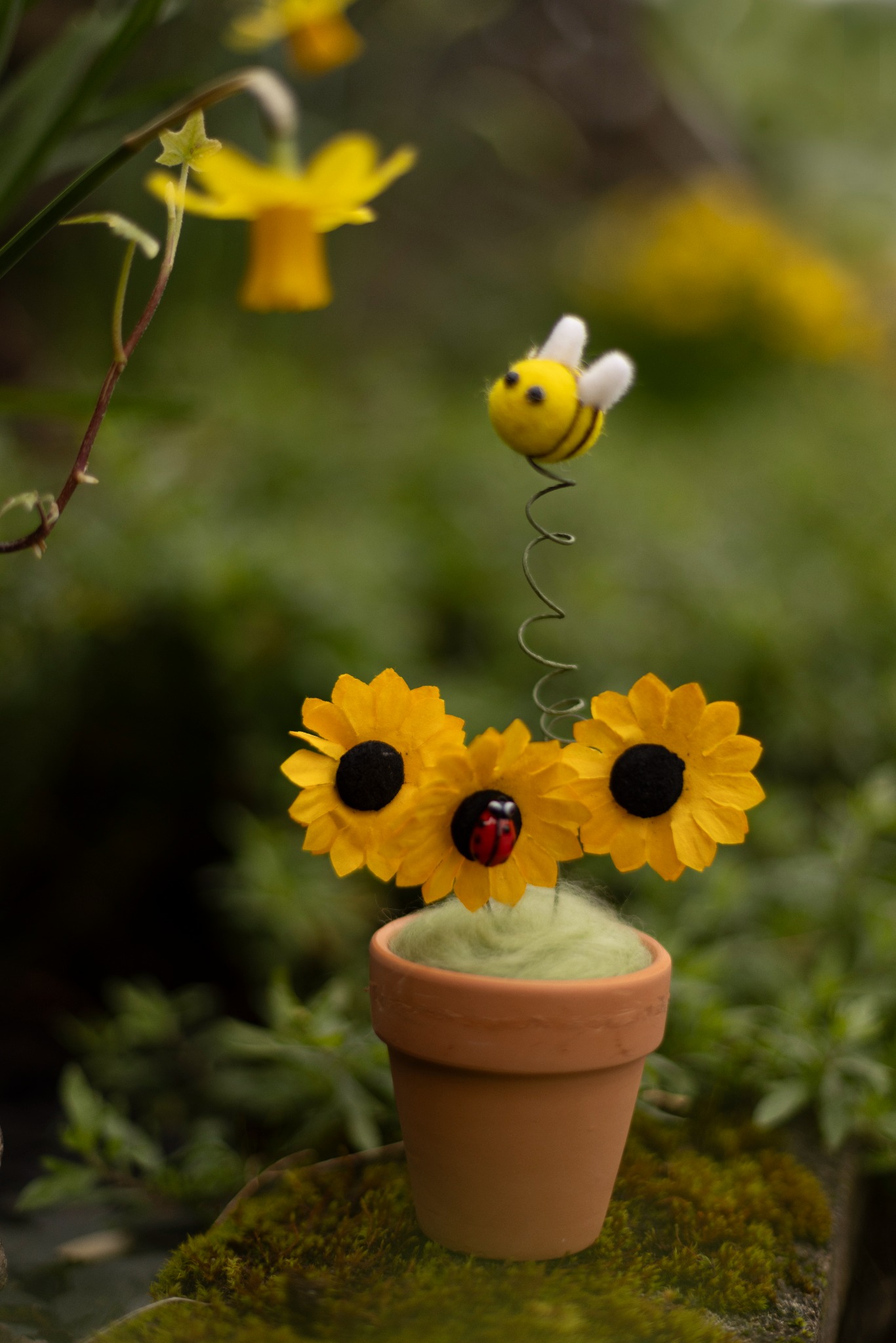 Needle-felted bee above yellow flowers in a small terracotta pot.
